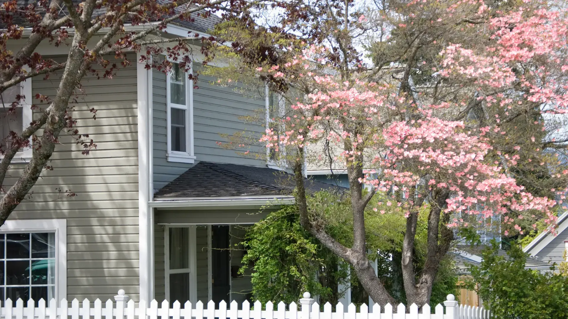 Two-story house with beige siding and spring blossoms against a white picket fence.